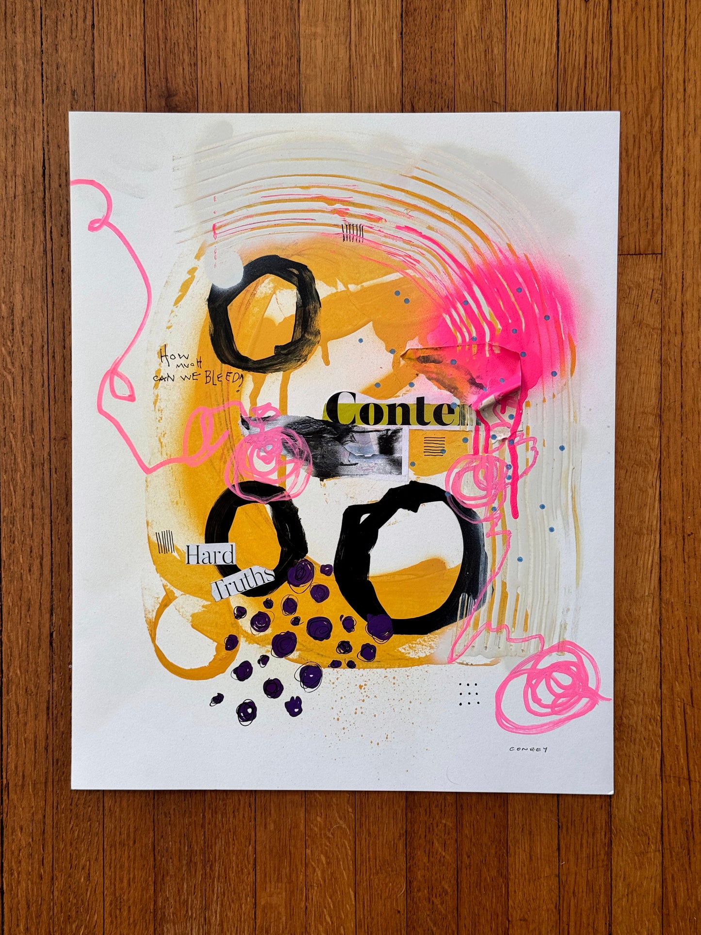 An abstract mixed-media art print featuring bold black circular shapes, vibrant yellow and orange paint drips, hot pink scribbles, and collage elements, displayed flat on a brown wooden surface.