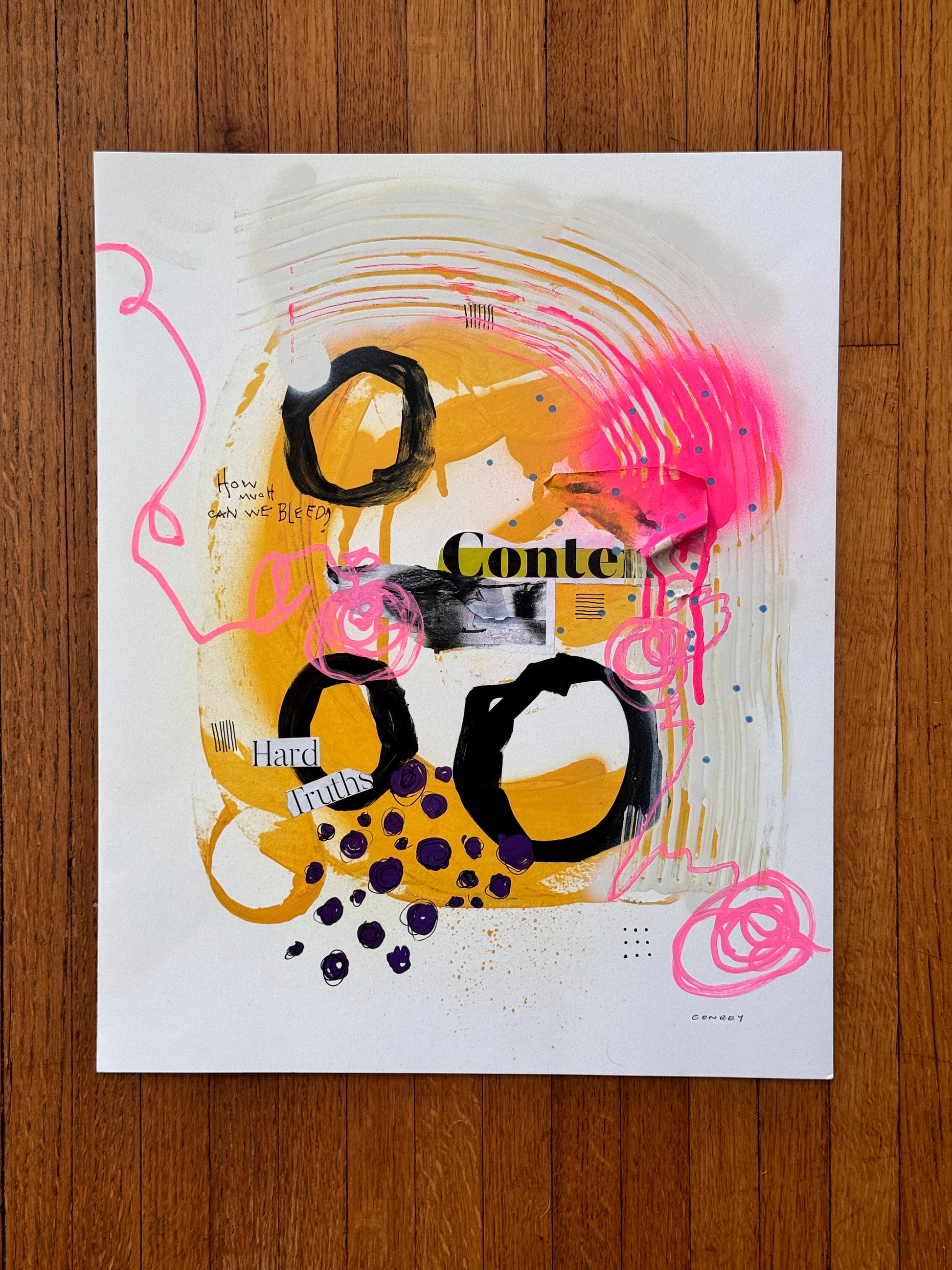 An abstract mixed-media art print featuring bold black circular shapes, vibrant yellow and orange paint drips, hot pink scribbles, and collage elements, displayed flat on a brown wooden surface.