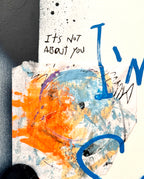 Abstract mixed-media art print detail featuring a textured white background with blue and orange paint splatters, a hand-drawn smiley face, and handwritten text reading 'It's Not About You'.