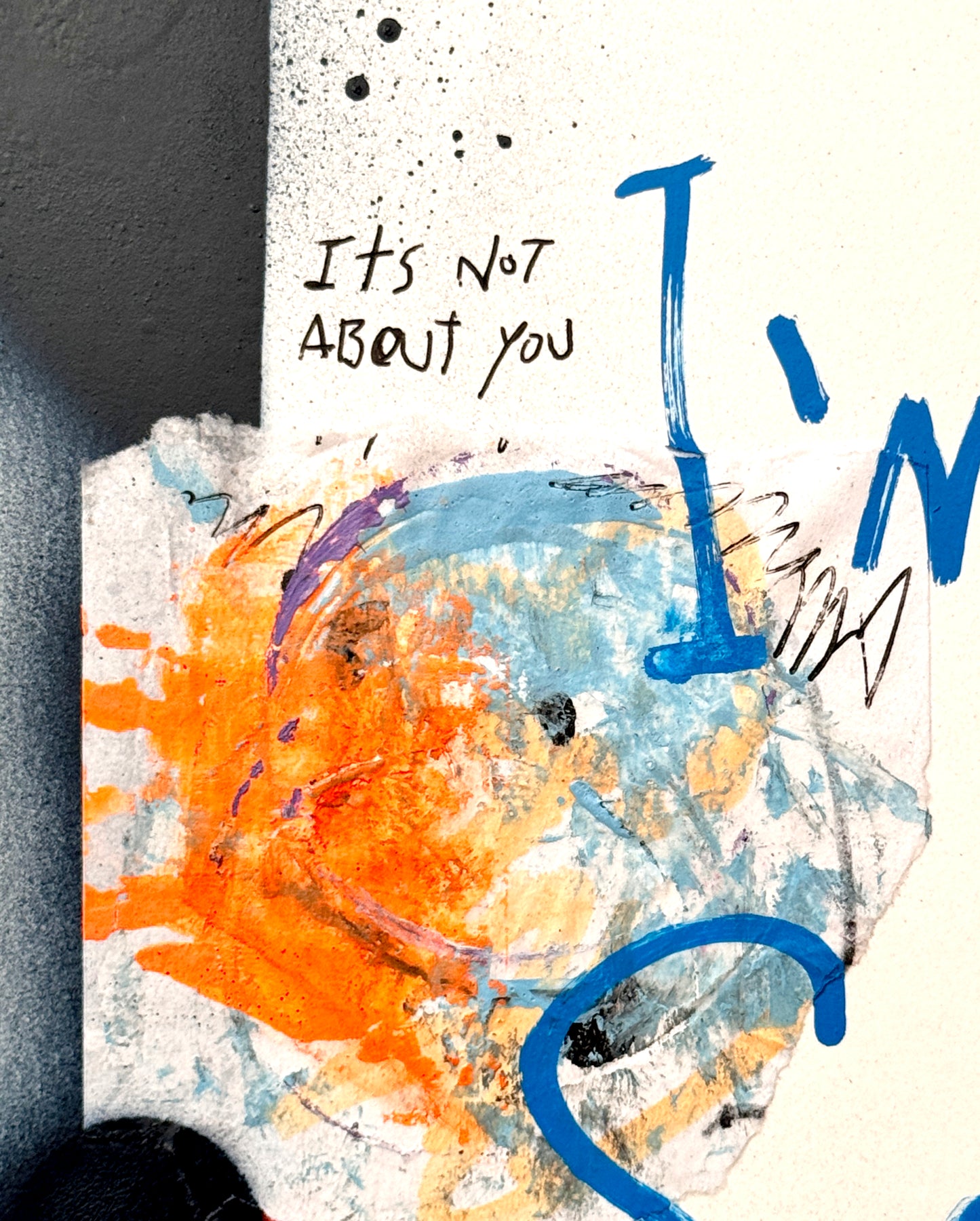 Abstract mixed-media art print detail featuring a textured white background with blue and orange paint splatters, a hand-drawn smiley face, and handwritten text reading 'It's Not About You'.