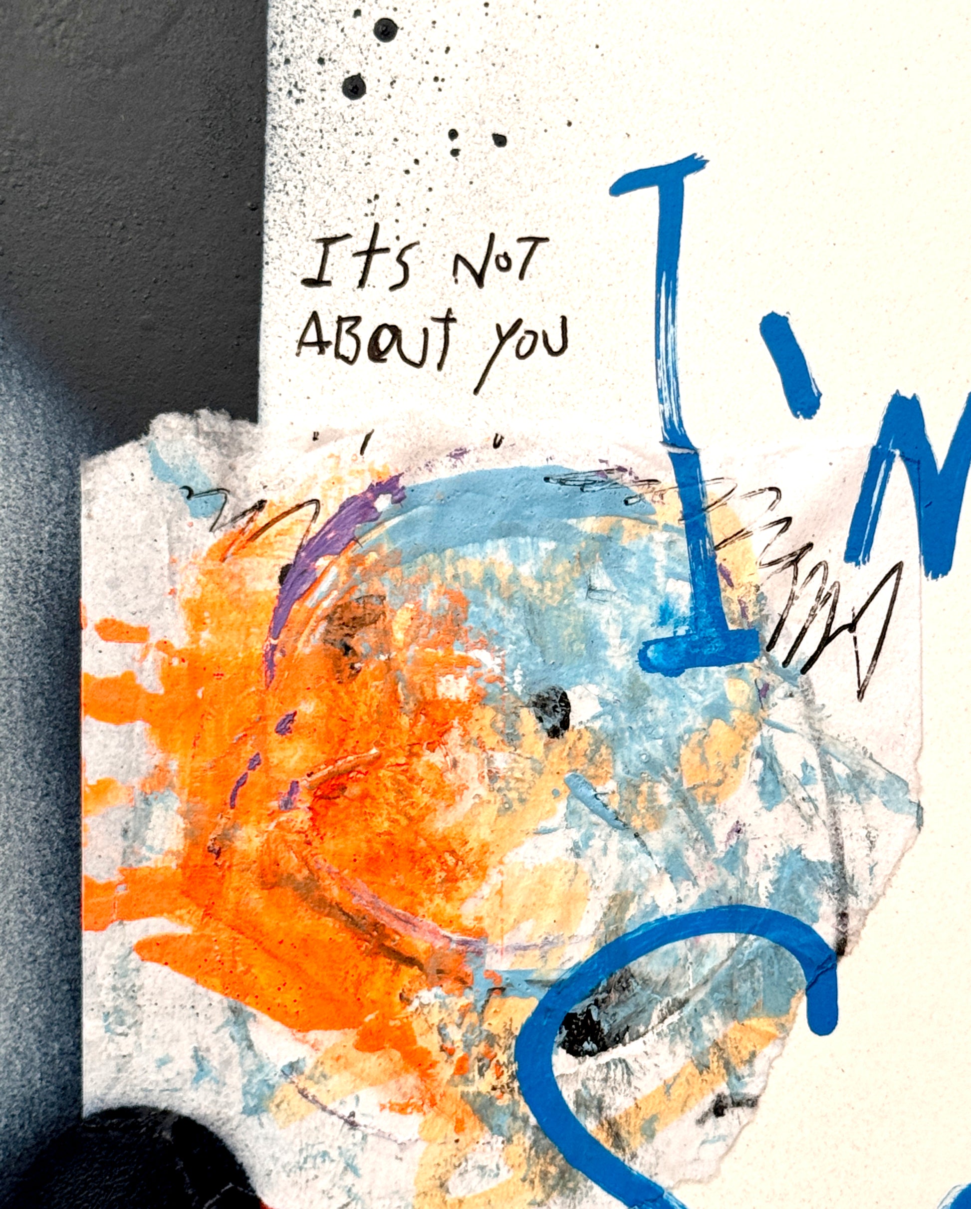 Abstract mixed-media art print detail featuring a textured white background with blue and orange paint splatters, a hand-drawn smiley face, and handwritten text reading 'It's Not About You'.
