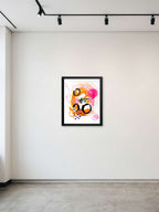 A framed abstract art print with vibrant orange, pink, and black brushstrokes on a white background, displayed on a minimalist gallery wall under track lighting.