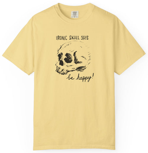 A pale yellow short-sleeve t-shirt with a black graphic of a skull and the text 'IRONIC SKULL SAYS be happy!' printed on the front, displayed against a plain white background.